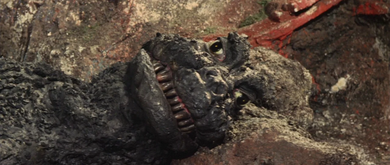 Son of Godzilla (1967) movie still: closeup — A dark, charred reptilian creature lies resting against a rugged, uneven stone surface. The ground…