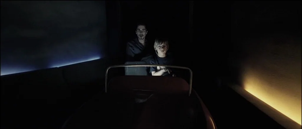 The Machinist (2004) movie still: medium — A man and a boy sit inside a dark vehicle on an indoor ride. The vehicle moves through a space with…
