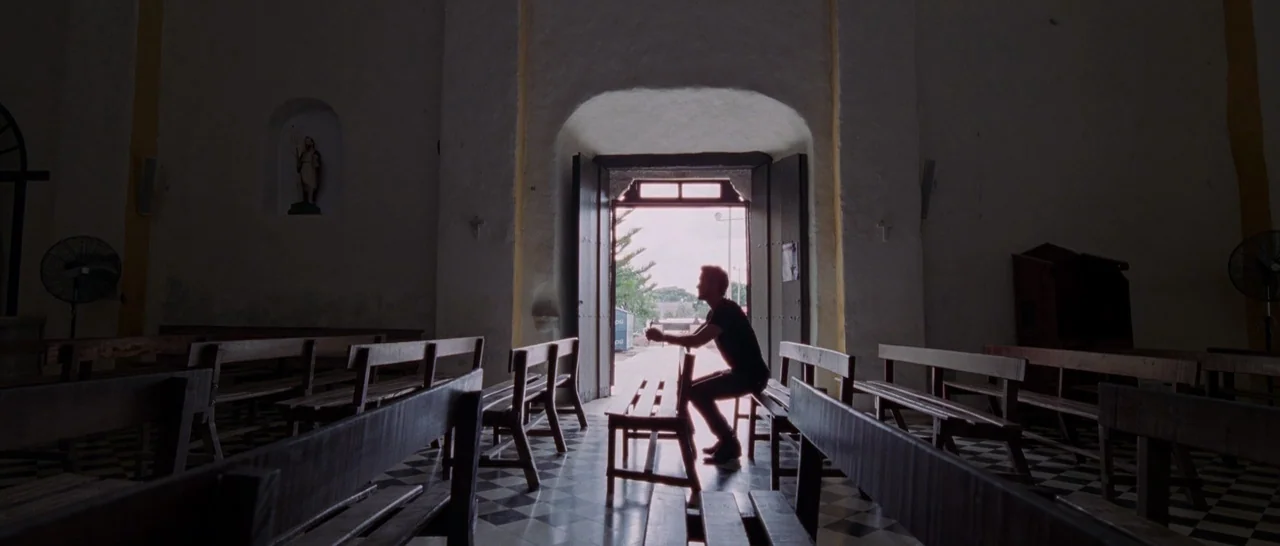 Song to Song (2017) movie still: wide — A sparsely furnished white-walled church with rows of wooden benches on a checkered floor. A man sits alone…