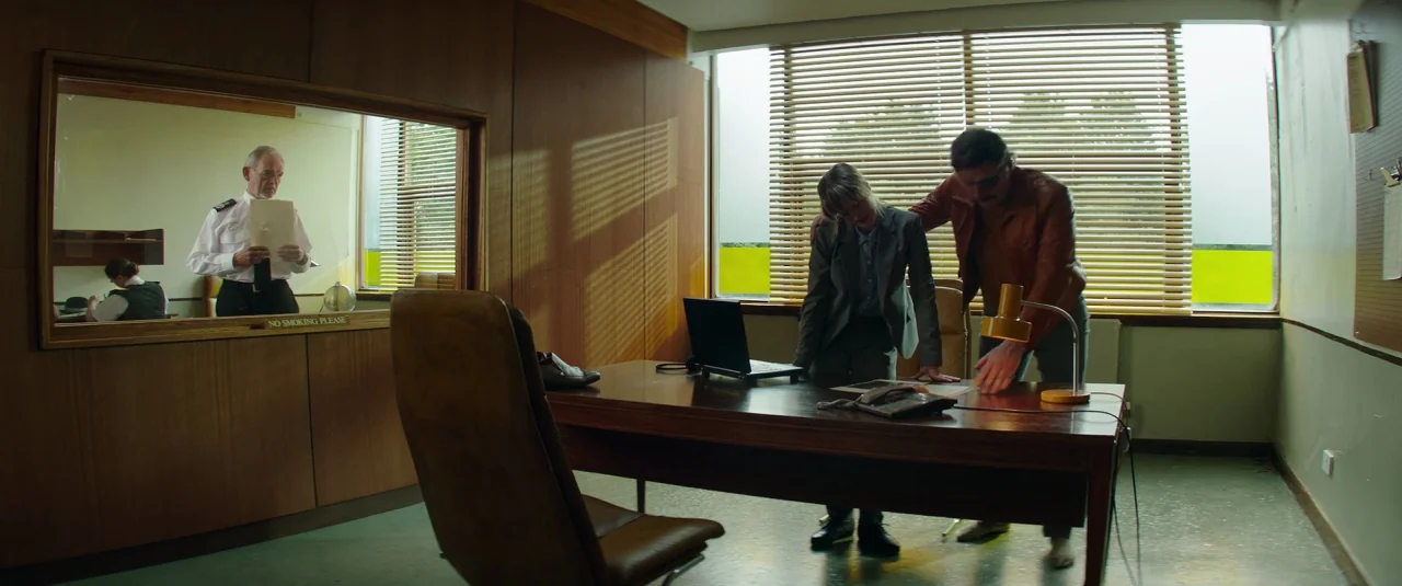 Mindhorn (2016) movie still: wide — A wood-paneled office contains a large desk and a window covered with horizontal blinds. A man and a woman stand…