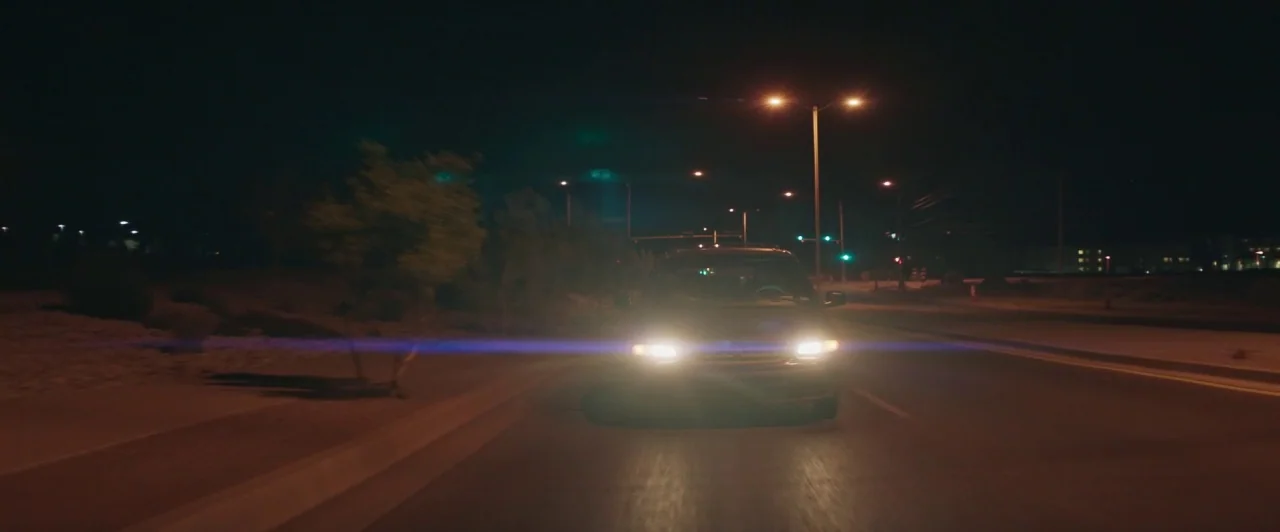 The Long Dumb Road (2018) movie still: wide — A dark asphalt road at night stretches forward under a row of orange streetlamps. An oncoming car with…
