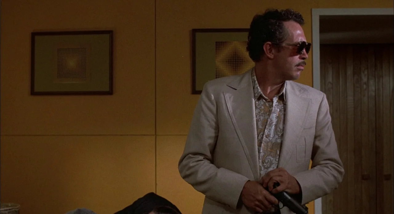 Bring Me The Head of Alfredo Garcia (1974) movie still: medium — A yellow-walled room features two geometric framed prints hanging behind a man. The…