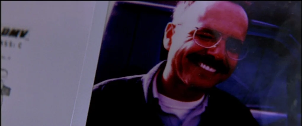 Memento (2000) movie still: closeup — A close-up view shows a glossy photograph of a middle-aged man. The man in the photo wears glasses and smiles…