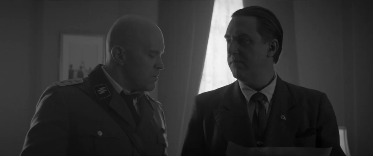 Bau, Artist at War (2025) movie still: medium — An interior office space with a window casting light across the room. Two men in historical uniforms…