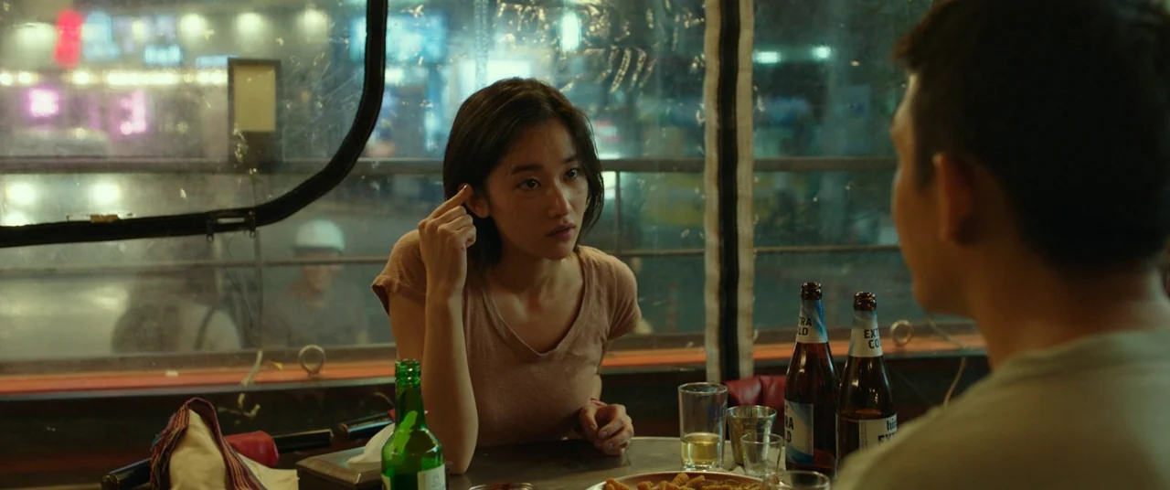 Burning (2018) movie still: medium — A small outdoor eating tent at night with a clear plastic wall overlooking city lights. A young woman sits at a…