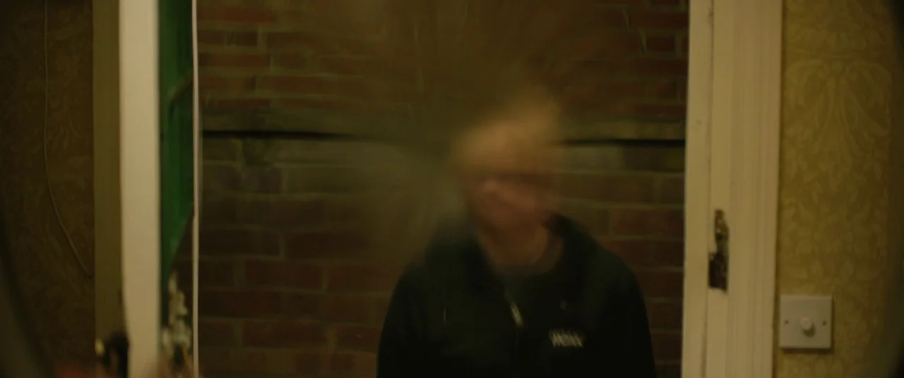 Yesterday (2019) movie still: medium — A dimly lit interior shows a person standing between a doorway frame. The subject's head is blurred as if…