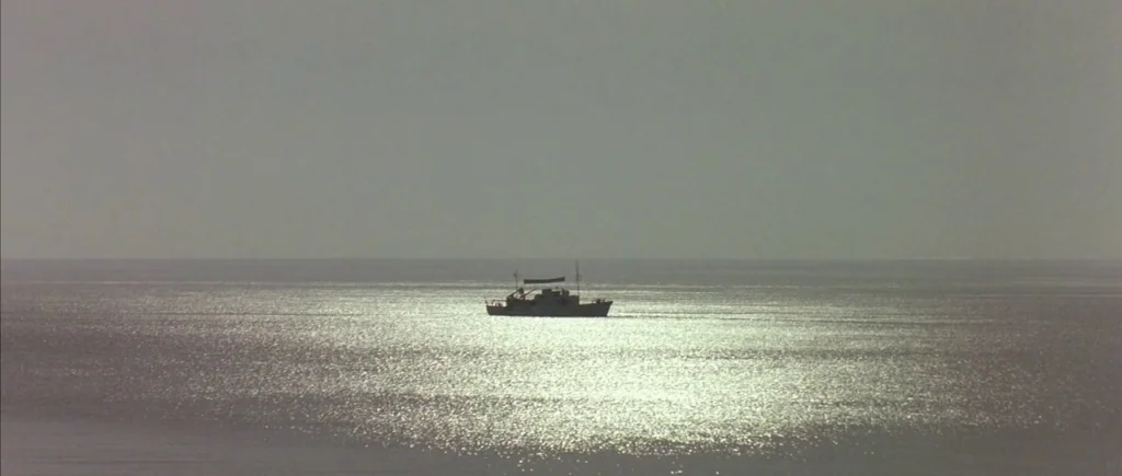 The Big Blue (1988) movie still: wide — A small vessel is centered in a vast, calm sea under a grey sky. The sun reflects brightly off the water…