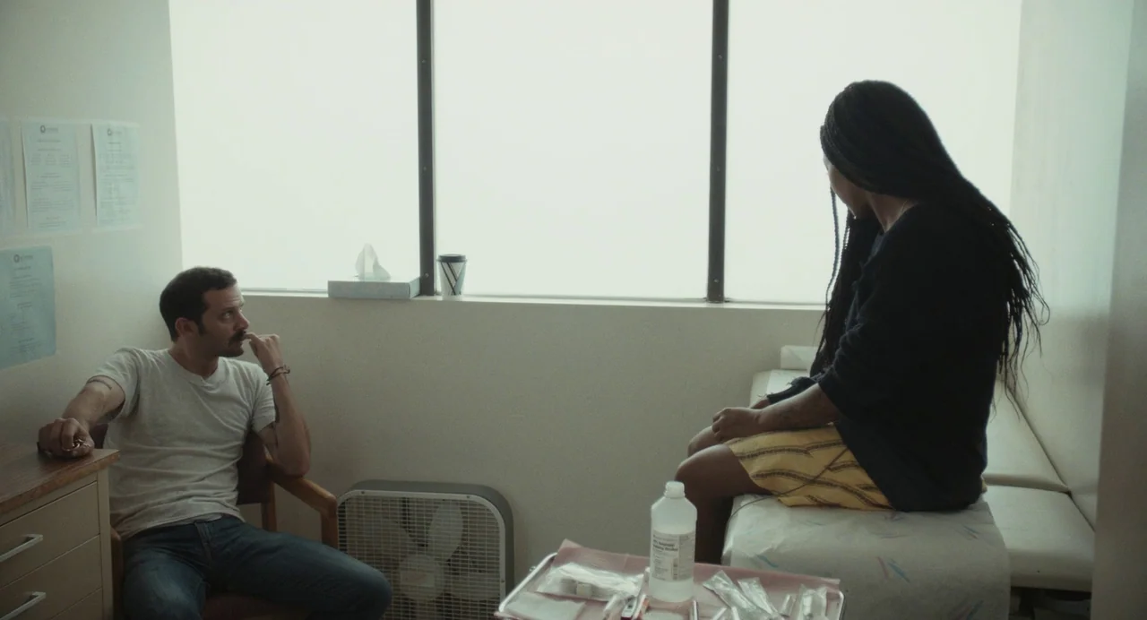 Test Pattern (2019) movie still: medium — An austere medical room features white walls, a patient bed, and documents posted prominently. Two…