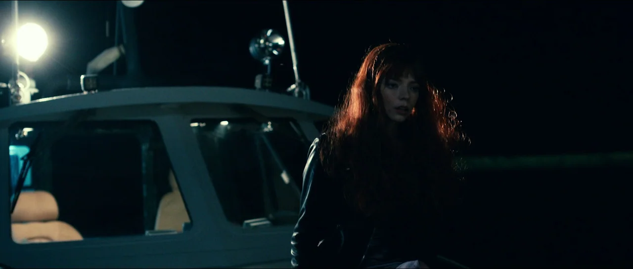 The Menu (2022) movie still: medium — A woman stands on the deck of a boat at night with darkness surrounding. She is positioned next to the pilot…