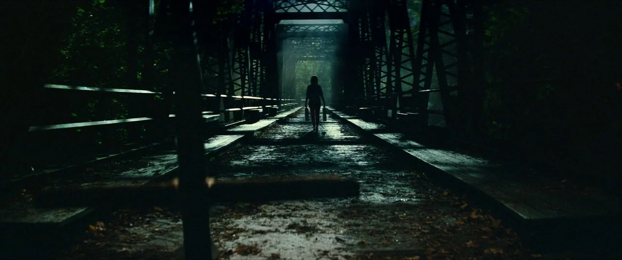 A Quiet Place Part II (2020) movie still: wide — A dark, wet metal truss bridge spans across a dense, wooded area. A lone figure walks away from the…