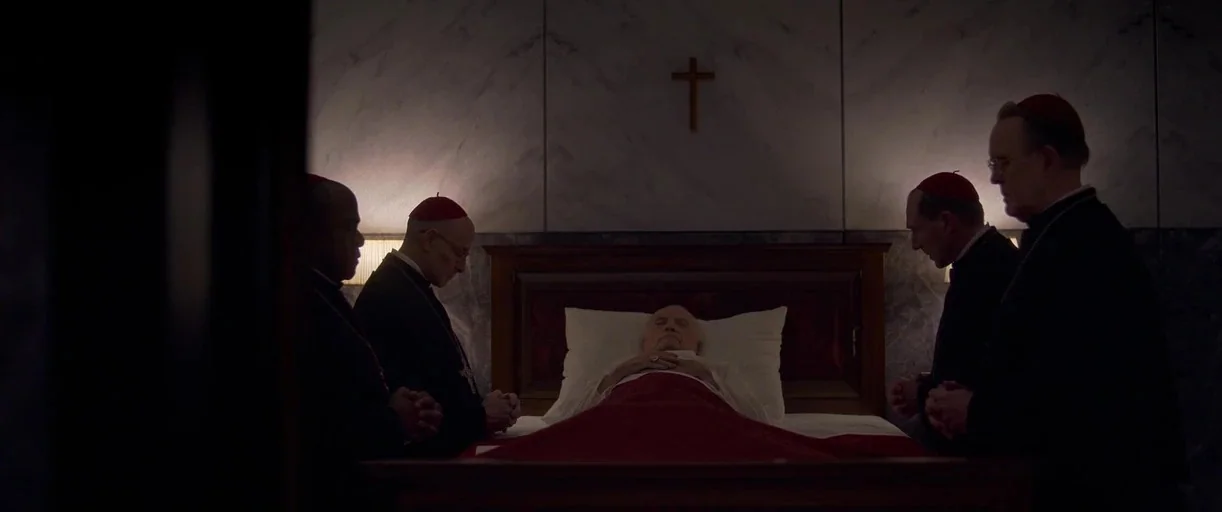 Conclave (2024) movie still: wide — A dimly lit room with gray marble walls and a small wooden wall crucifix. Four clergymen in black standing…