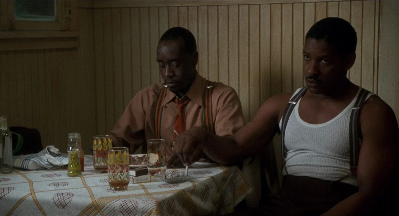 Devil In A Blue Dress (1995) movie still: medium — A small room with vertical wood paneling and a table covered by a patterned cloth. Two men sit at…