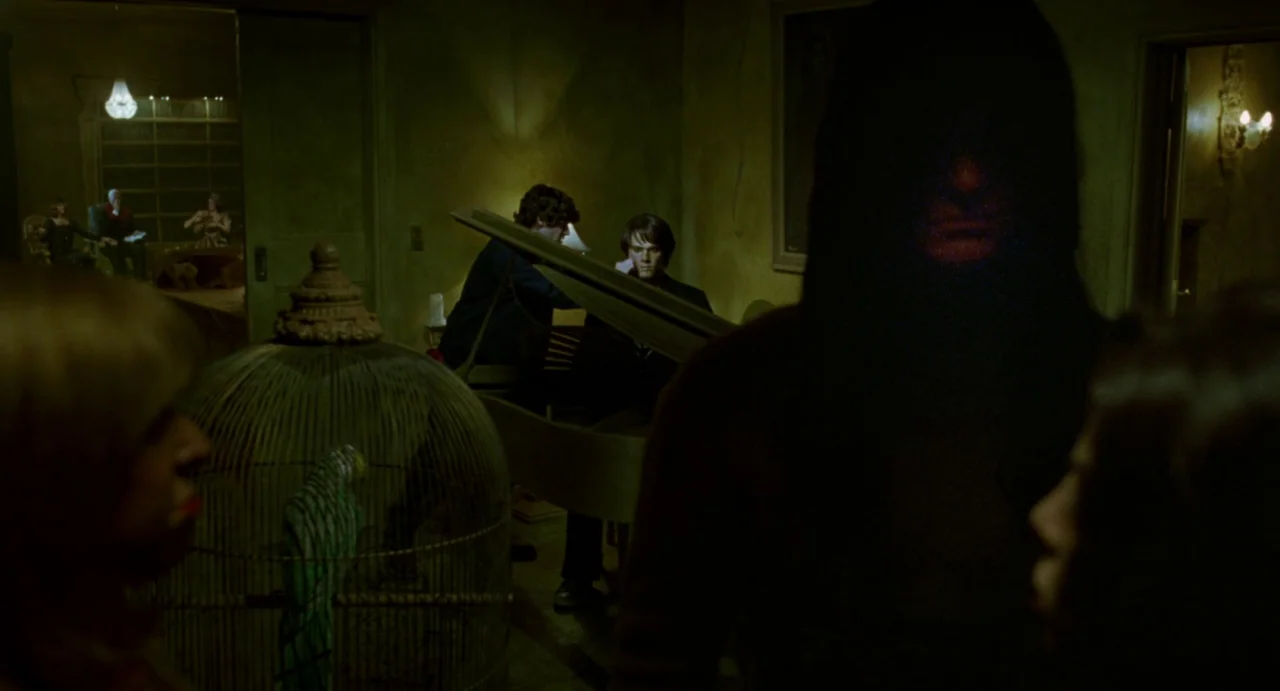 House of Wax (2005) movie still: medium — An dimly lit, olive-toned room contains a birdcage, a piano, and several people. A silhouetted figure…