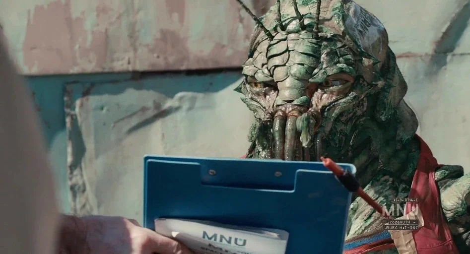 District 9 (2009) movie still: closeup — A weathered industrial setting with faded teal walls and metallic debris in the background. An alien being…