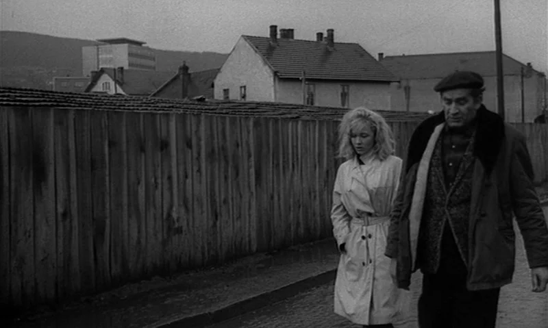 Man Is Not Bird (1965) movie still: fullbody — A woman and an older man walk along a concrete sidewalk beside a fence. Houses and buildings are…