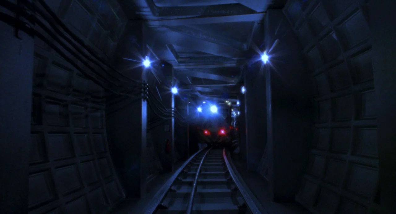 Resident Evil (2002) movie still: establishing — A dark subterranean subway tunnel features grid-patterned concrete walls and visible electrical…
