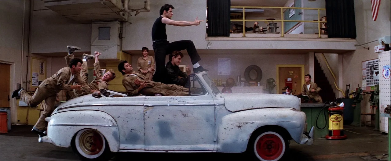 Grease (1978) movie still: wide — A cluttered auto shop contains a large, weathered, light-colored convertible as a centerpiece. Multiple young men…