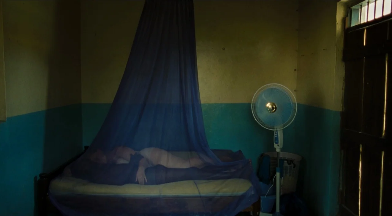 Paradise: Love (2012) movie still: wide — A spare interior space features two-toned walls painted in blue and yellow hues. A bed covered by a…