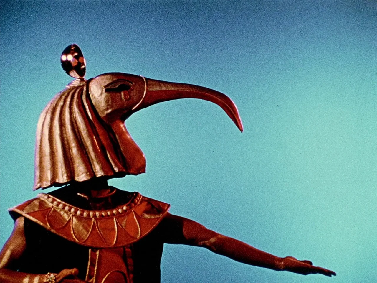 Space is the Place (1974) movie still: medium — A figure wears an elaborate metallic ibis mask against a flat blue screen. The person stands with…