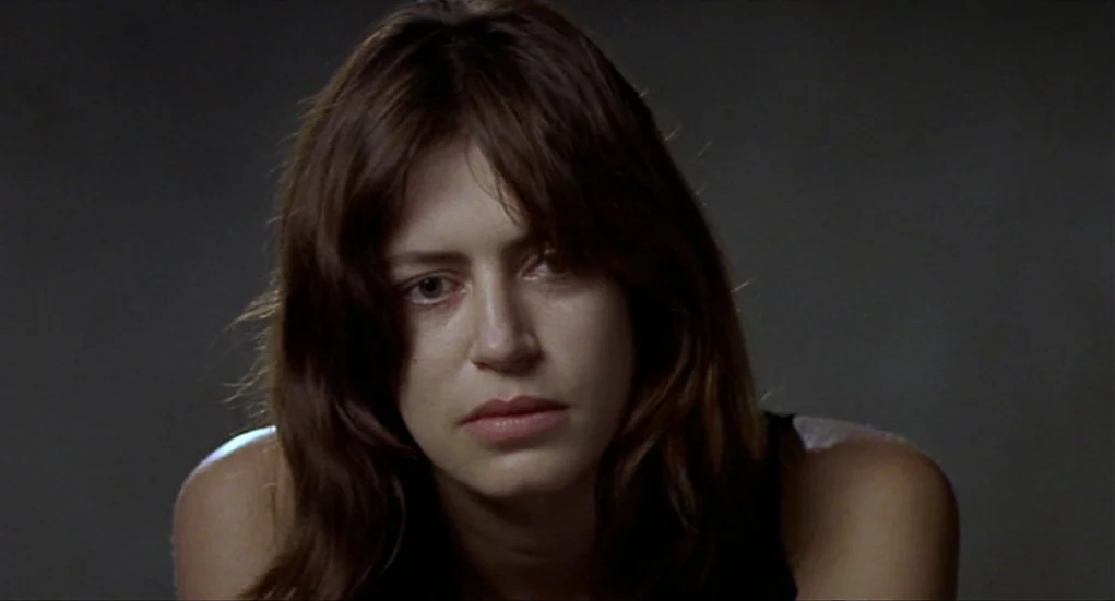 Tiresia (2003) movie still: closeup — A woman stands in front of a dark, plain studio background. She looks forward while her face is lit by soft,…