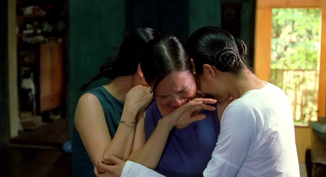 The Vertical Ray of the Sun (2000) movie still: medium — An interior space with cool dark wall tones and a bright doorway visible. Three women hold…