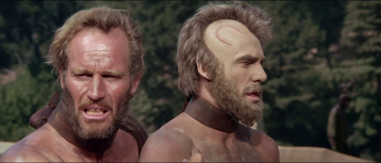 Planet of the Apes (1968) (1968) movie still: medium — Two bare-chested men stand side-by-side in a dense forest setting. Both subjects wear thick…