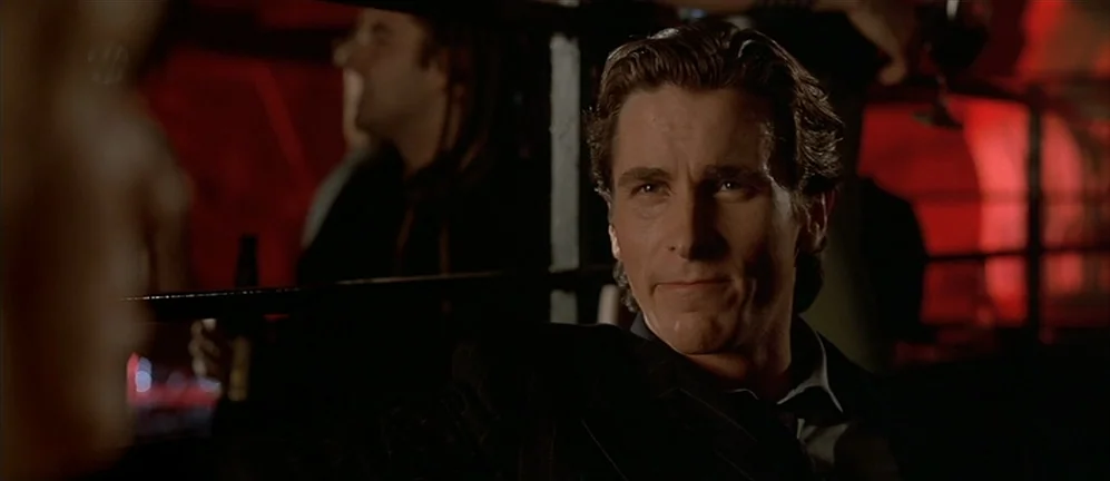 American Psycho (2000) movie still: medium — A dark, crowded nightclub interior is filled with red artificial light and patrons. A man sits at a…