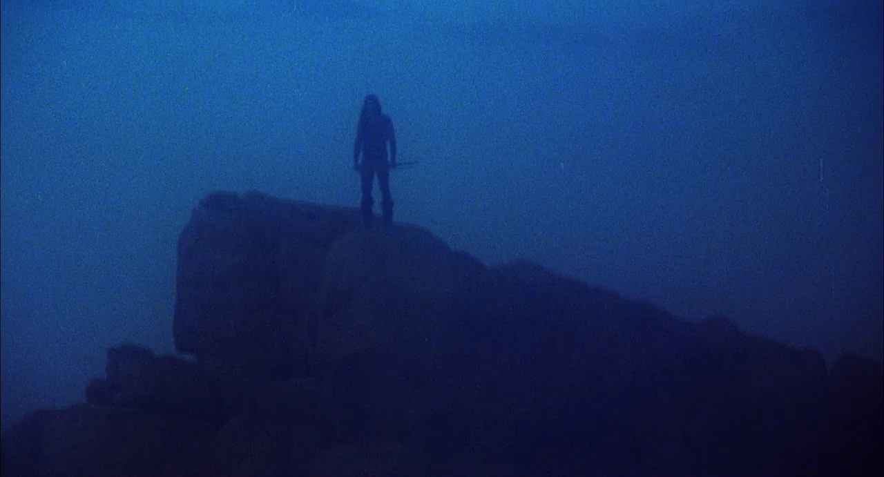Conquest (1983) movie still: wide — A dark rocky outcrop is shrouded in thick blue mist at night. A lone figure stands atop the rock formation…
