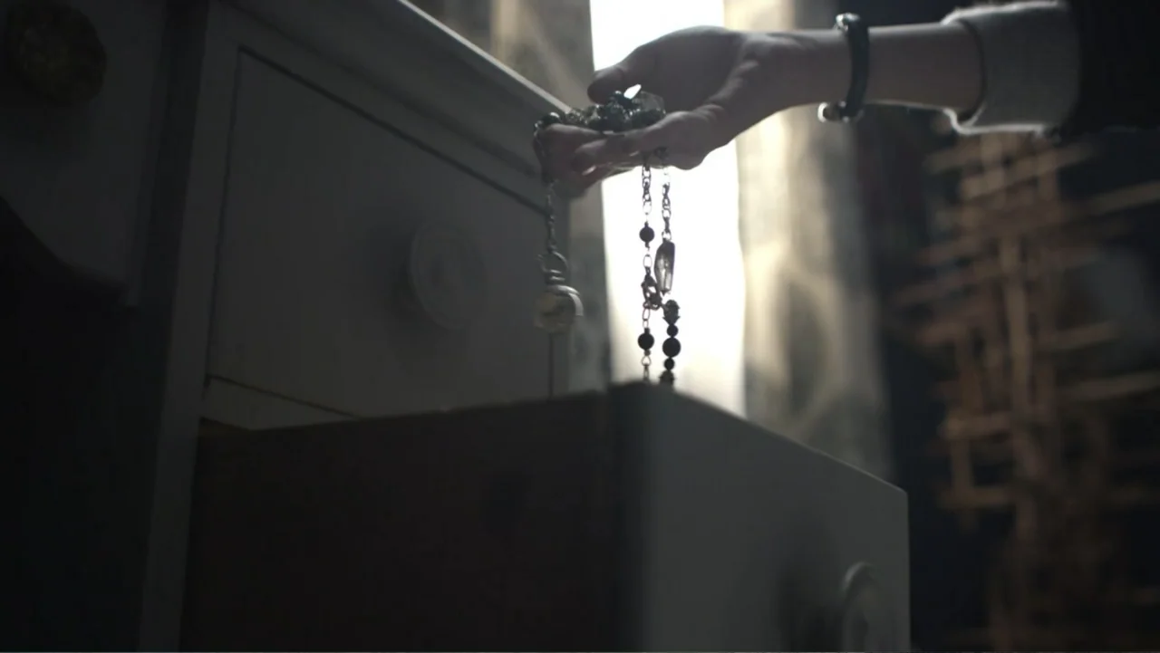 Totem (2022) movie still: closeup — A wooden dresser with an open drawer sits in front of a bright window. A hand holds a beaded necklace above the…
