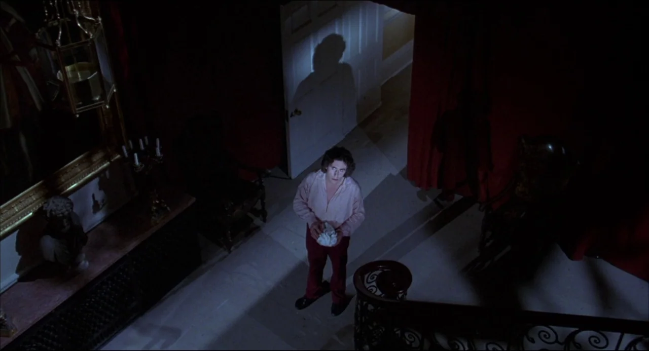 Gothic (1986) movie still: wide — A dark foyer features red curtains, a bust statue, and an intricate iron staircase. A man stands on the landing…