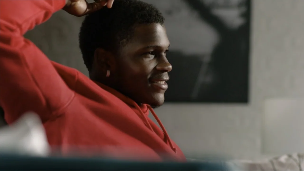 We Beat the Dream Team (2025) movie still: medium — A living room interior features a white brick wall and a framed picture. A young man in a red…