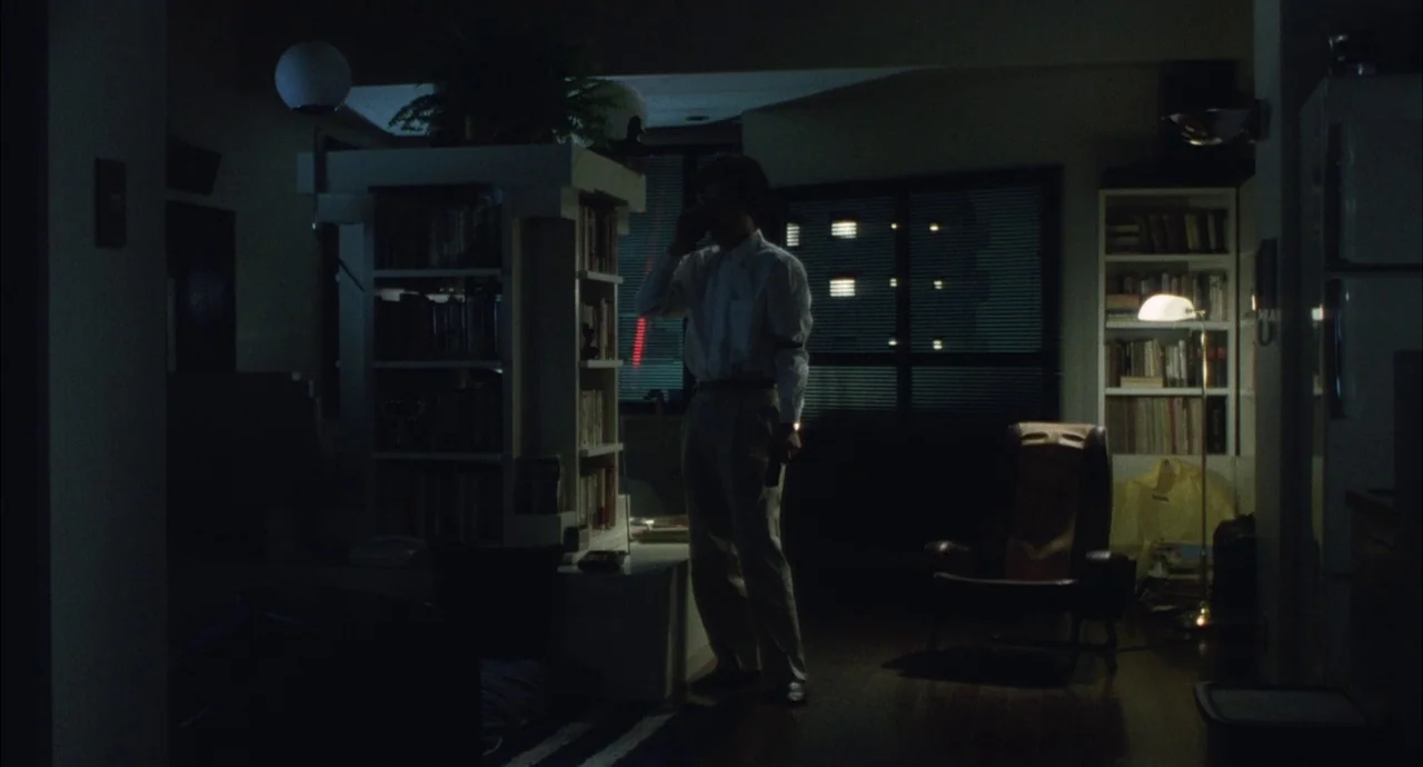 The Discarnates (1988) movie still: wide — A dimly lit room contains a white modular bookshelf and an armchair. A man in a white shirt stands in the…