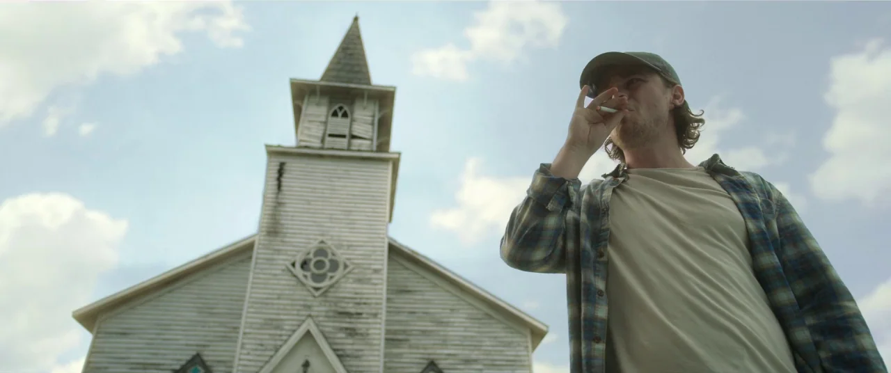 In The Tall Grass (2019) movie still: medium — A white clapboard church stands under a cloudy sky. A man in a baseball cap smokes a cigarette while…