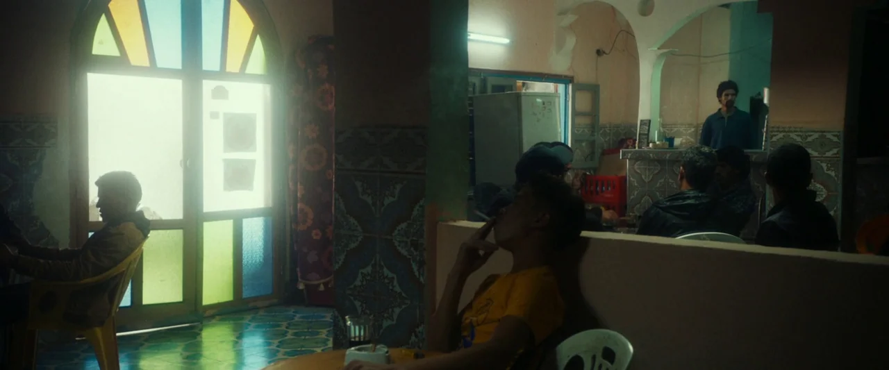 Animalia (2023) movie still: wide — A dimly lit room features tiled walls, a stained glass door, and yellow plastic chairs. Several young men sit at…