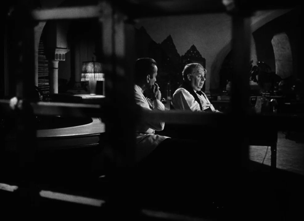 Casablanca (1942) movie still: medium — A dark, dimly lit study features architectural arched elements and a table lamp. Two men sit in a…
