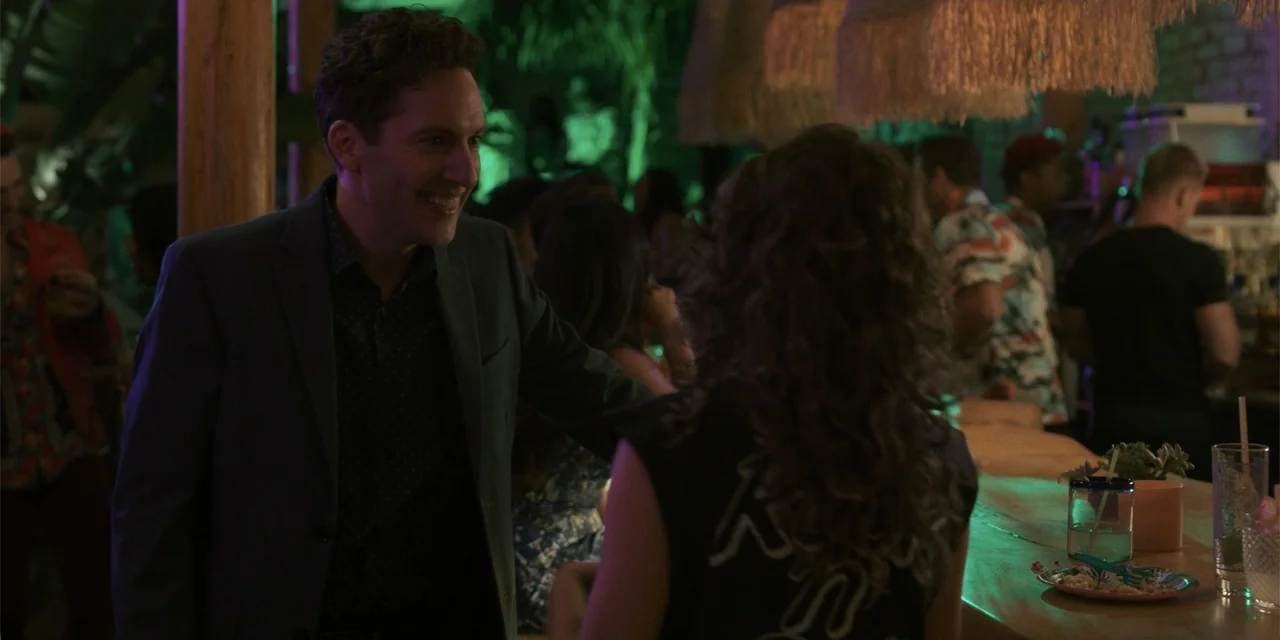 Sitting in Bars with Cake (2023) movie still: medium — A dimly lit bar at night featuring wooden counters and hanging straw decorations. A man in a…