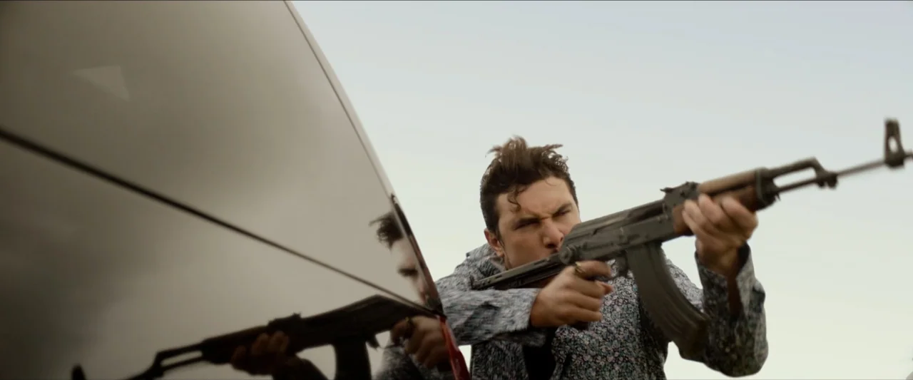 The Marksman (2021) movie still: medium — An open desert location under a bright, overcast sky during the day. A man crouches near a reflective…