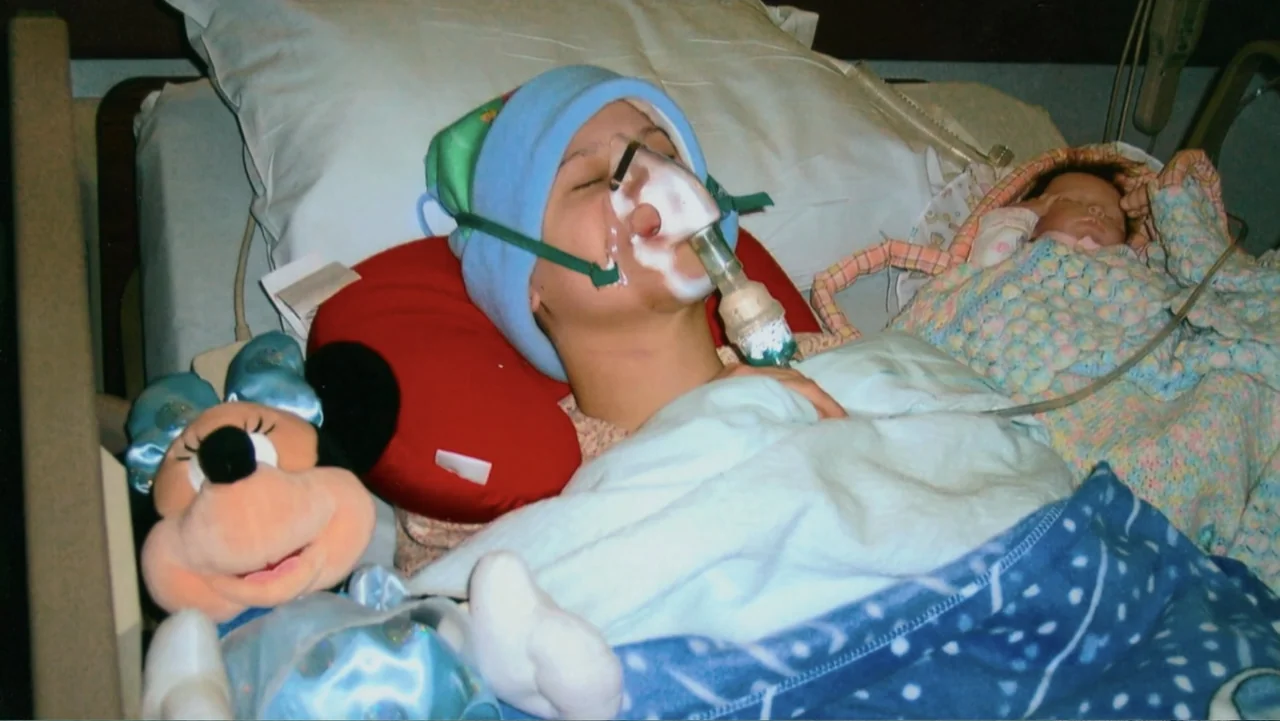Mommy Dead and Dearest (2017) movie still: medium — A hospital bed with white sheets holds a child wearing a medical oxygen mask. A Minnie Mouse…