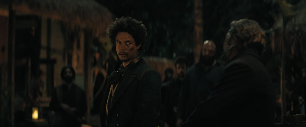 The Bluff (2026) movie still: medium — A nighttime outdoor scene features a gathering of people in a dimly lit village area. A man with a dark afro…
