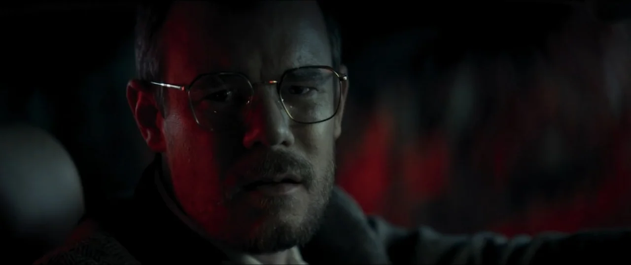Offseason (2022) movie still: closeup — Inside a dark vehicle at night, the interior is lit by red ambient light. A man in eyeglasses focuses on the…