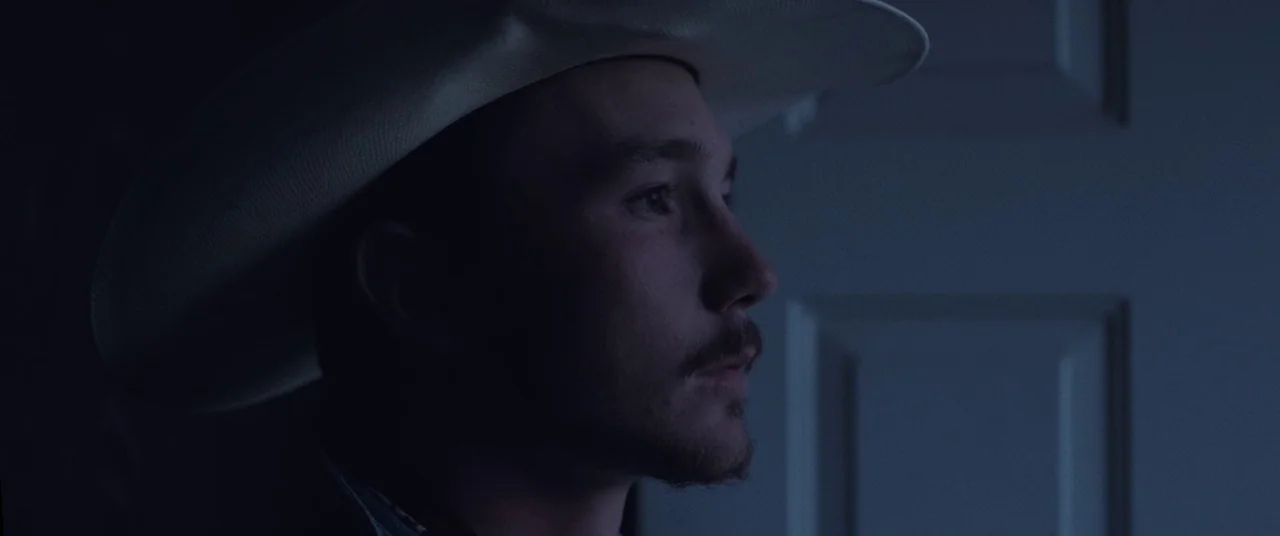 The Rider (2017) movie still: closeup — A man stands in a dimly lit room against a plain white interior door. The subject wears a cowboy hat while…