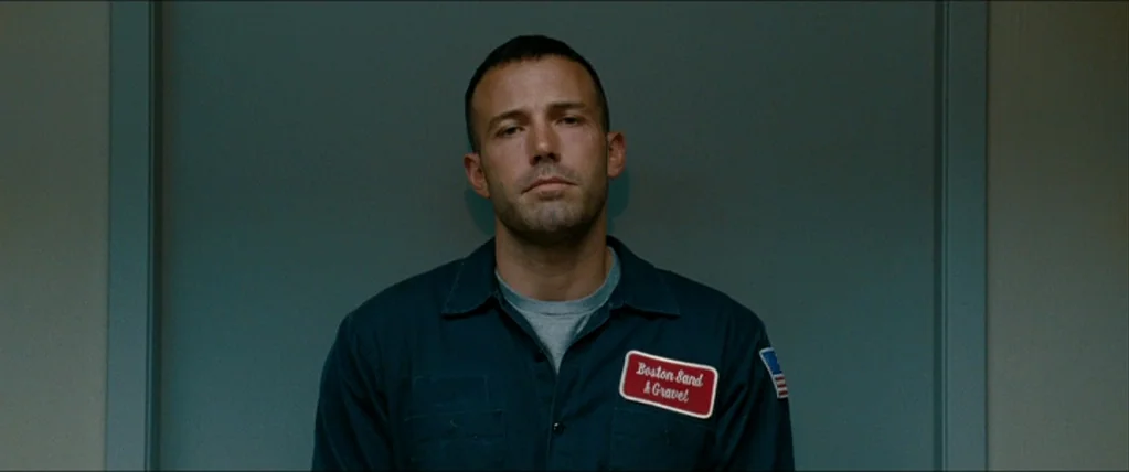 The Town (2010) movie still: medium — A man stands before a plain blue-grey wall in what resembles a booking room. He is dressed in a dark uniform…
