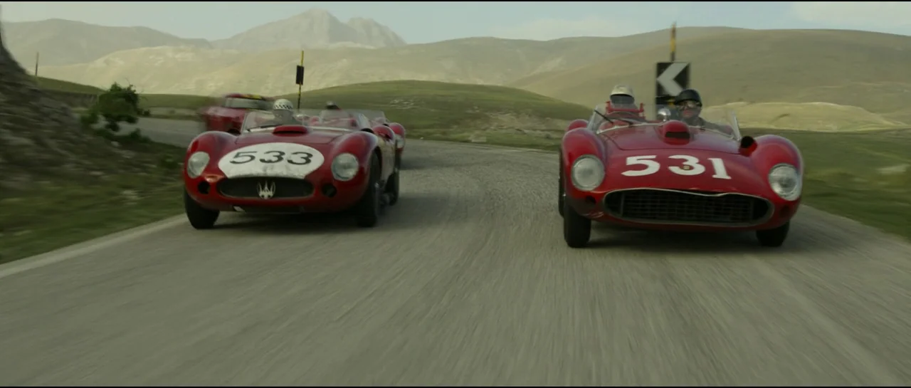 Ferrari (2023) movie still: wide — Two vintage red race cars drive side by side on a winding mountain road. Rolling green hills and distant peaks…