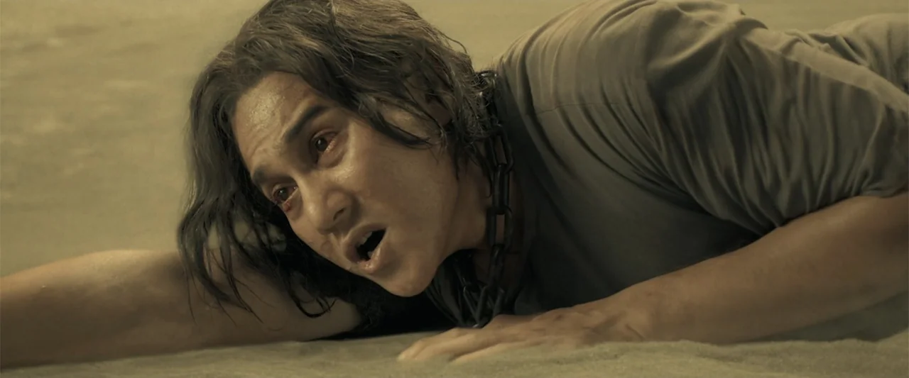 The Unholy (2022) movie still: closeup — A bare, dusty interior floor with a man lying flat on his stomach. A heavy metal chain is wrapped around…