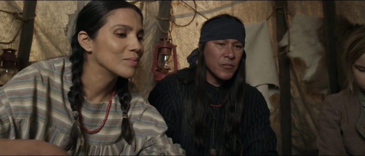 Dead for a Dollar (2022) movie still: medium — A rustic interior constructed from hide and poles contains two people. A woman and a man sit side by…