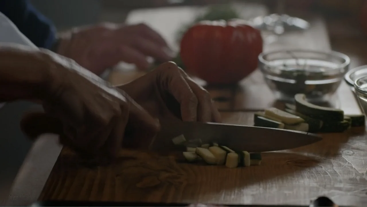 A Pinch of Portugal (2023) movie still: closeup — A close view of hands working on a wooden cutting board in a home kitchen. The individual slices…