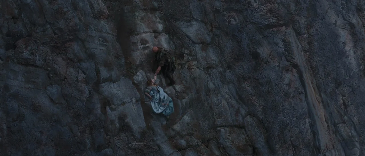 Tale of Tales (2015) movie still: wide — A vertical rock cliff face with uneven textures occupies the frame. A man reaches down to assist a woman in…