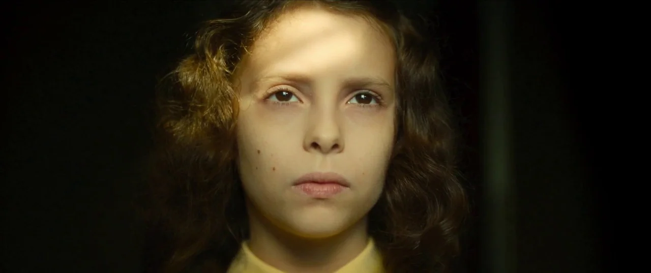 Earwig (2022) movie still: closeup — A dark indoor space provides contrast for a close shot of a young girl. The child stares forward while light…