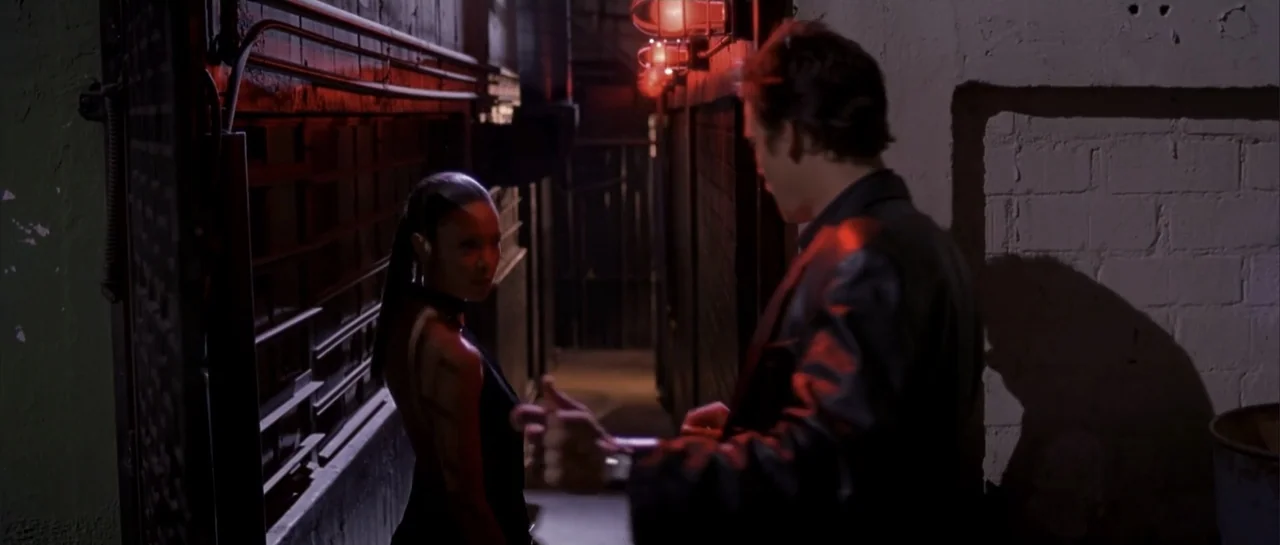 Shade (2003) movie still: medium — A narrow outdoor passage features dark brick walls and red neon warning lights. A man in a leather jacket stands…