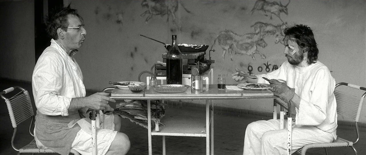 The Last Battle (1983) movie still: wide — An austere room features white walls decorated with primitive charcoal drawings of animals. Two men in…